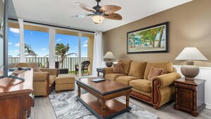 A spacious living room with a large sofa, a coffee table, and a ceiling fan. There is a large window with a view of palm trees and a balcony. - Beach Condo/ Island Beach Resort/No Cleaning Fee/ Condo 308 (Jensen Beach)