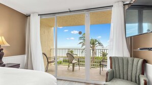 A hotel room with a bed, a chair, a table, and a balcony with a view of palm trees and a clear sky. - Beach Condo/ Island Beach Resort/No Cleaning Fee/ Condo 308 (Jensen Beach)