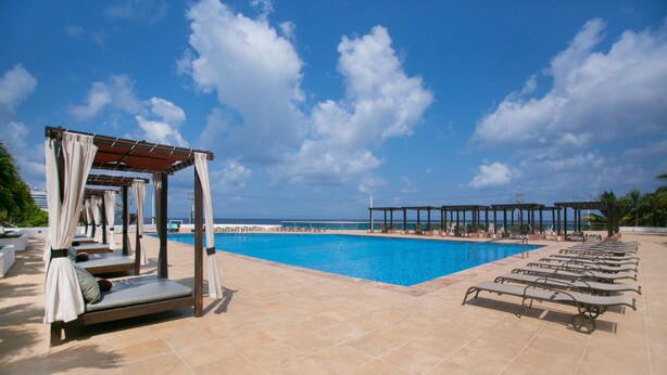 Un espace piscine doté de chaises longues et d’une pergola agrémentée de rideaux, offrant une vue dégagée sur un ciel bleu azur et une perspective lointaine sur l’océan.