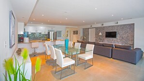 Un salon moderne avec une table à manger en verre, des chaises blanches, un canapé gris et un téléviseur à écran plat fixé à un mur en pierre.
