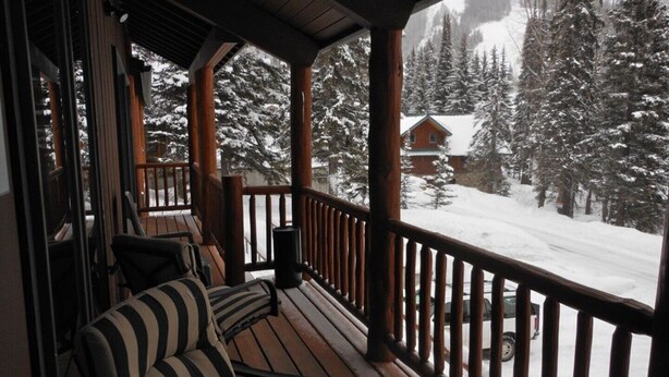 Une terrasse en bois avec un canapé et une rambarde, donnant sur un paysage enneigé parsemé d’arbres et sur une cabane.
