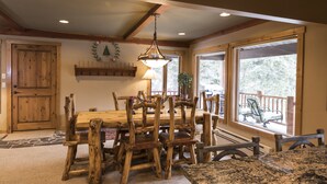Une table à manger en bois, des chaises, un luminaire suspendu et de grandes fenêtres donnant sur l’extérieur.