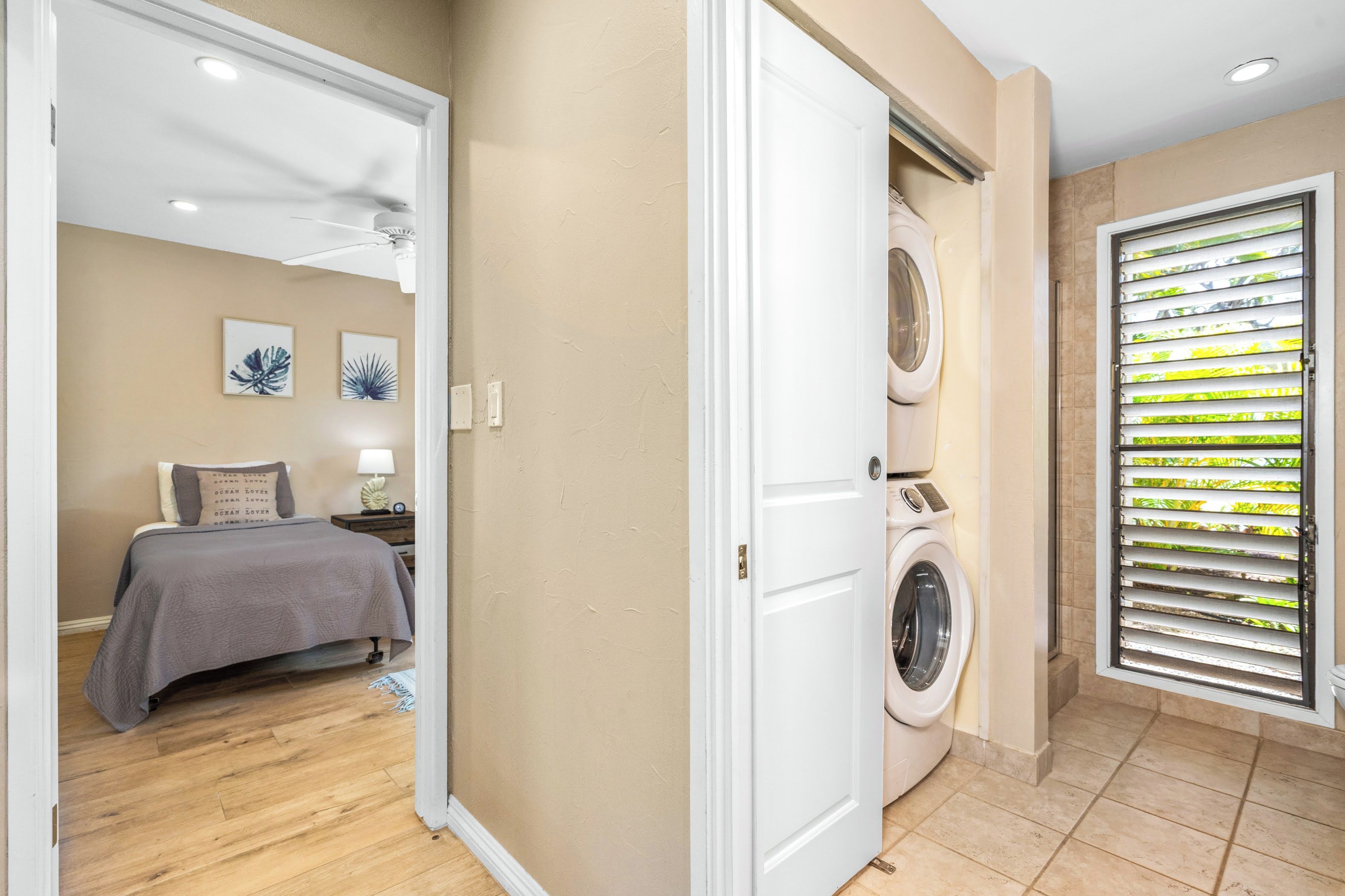 Ein Waschraum mit Waschmaschine und Trockner, eine Tür zu einem Schlafzimmer mit Bett und Nachttisch sowie ein Fenster mit Jalousien.