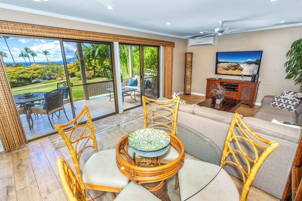 A spacious living area with a large sliding glass door leading to a balcony with a view of a lush garden and the ocean. There is a comfortable seating area with a sofa and two wicker chairs, a television on a wooden stand, and a small table with a decorative bowl.
