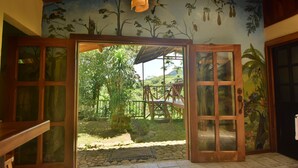 Quarto com uma porta grande aberta para área externa coberta, com deck de madeira e um mural com cenas da natureza na parede.