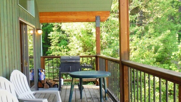 A wooden deck with white plastic chairs and a table, overlooking a forested area. - Comfortable & Spacious Ski-In/Ski-Out Cabin (Whitefish)