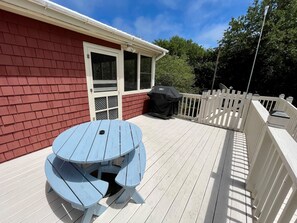 Une terrasse couverte avec une table de pique-nique et un barbecue, entourée d’une clôture blanche et d’un bâtiment rouge.