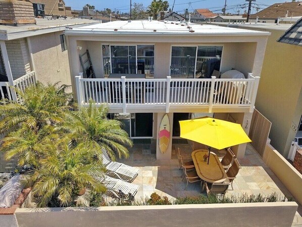 A two-story house with a balcony, a patio with chairs and a table, and a surfboard.