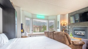 A hotel room with a large bed, a brown sofa, a fireplace, a TV, and a view of mountains through the windows. - Studio that sleeps 4 w/stunning mountain views, hot tub, free wifi, & parking (Copper Mountain)