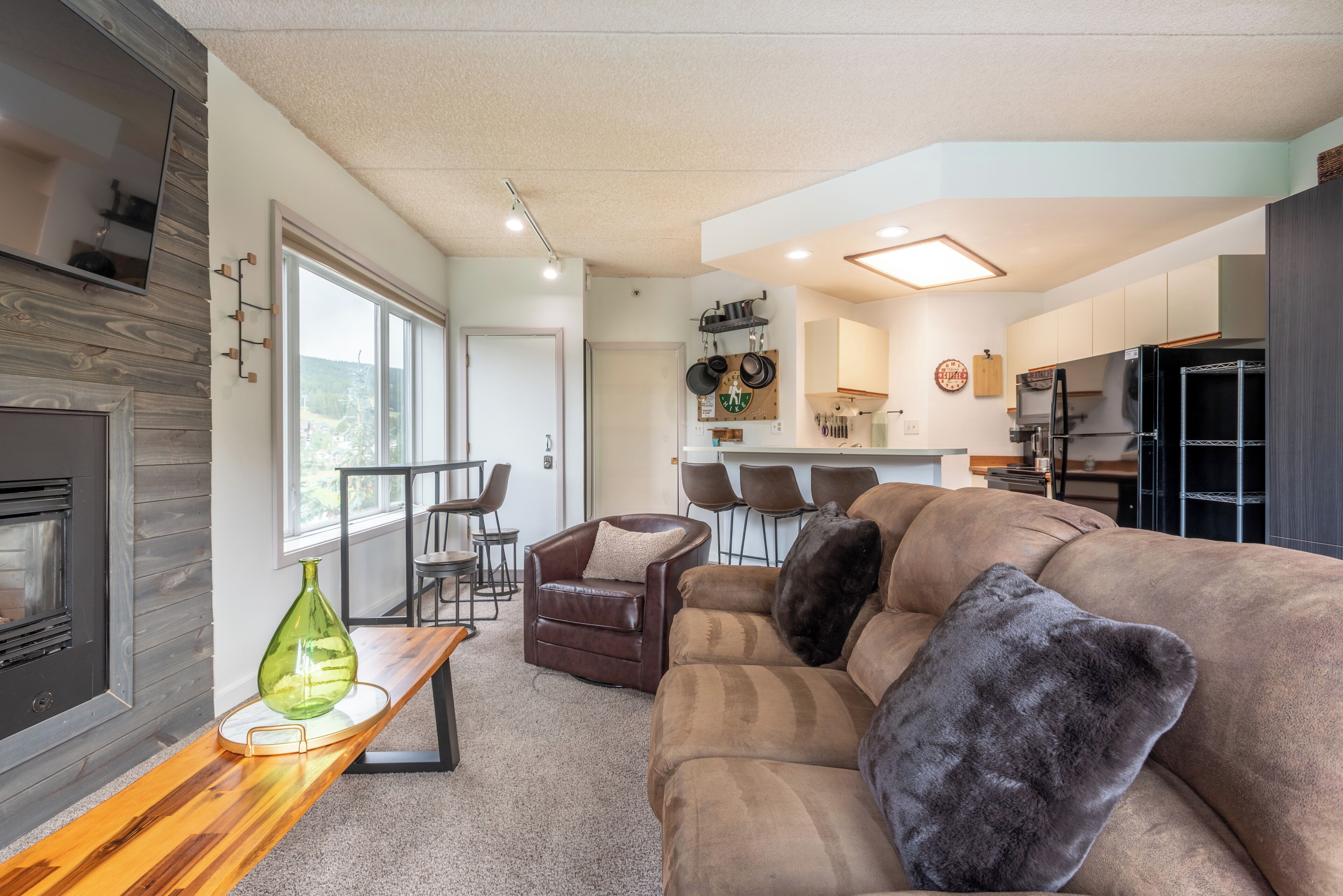 A modern living room with a brown sofa, a wooden coffee table, and a TV mounted on the wall. There is a kitchen area with a refrigerator, stove, and bar stools.