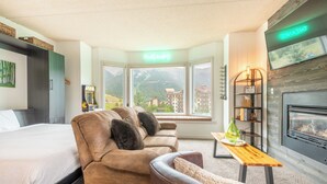 A hotel room with a bed, two sofas, a wooden table, a fireplace, a TV, and a large window with a view of mountains. - Studio that sleeps 4 w/stunning mountain views, hot tub, free wifi, & parking (Copper Mountain)