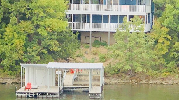 Une maison à deux étages avec une jetée qui s’avance dans un plan d’eau, le tout entouré d’une végétation luxuriante.