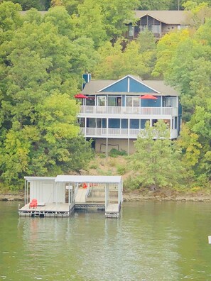 A two-story house with a dock extending into a body of water, surrounded by dense greenery. - Lakefront Margaritaville- Private Dock, 2 Decks, Firepit, Main Channel View (Osage Beach)