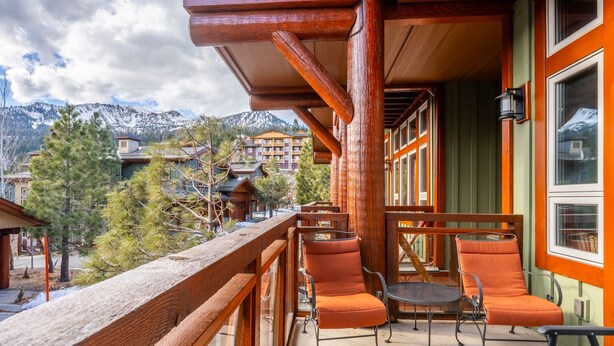 Ein Holzbalkon mit orangefarbenen Stühlen und einem Tisch, der auf eine gebirgige Landschaft mit Gebäuden blickt.