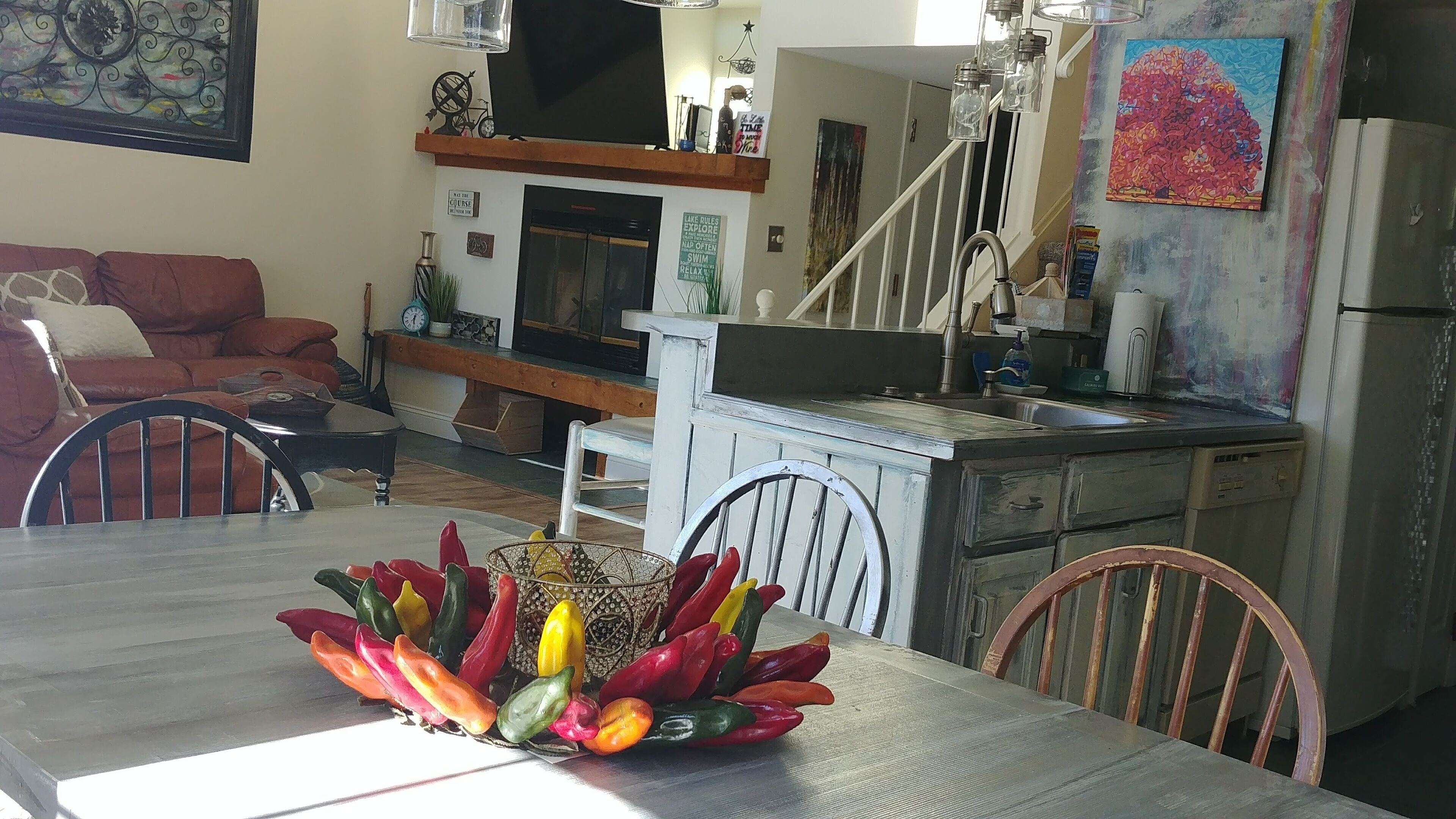 A dining area with a table set for two, a kitchen with a sink and cabinets, a staircase, and a living room with a sofa and a fireplace.