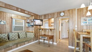 A living room with a wooden paneled wall, a sofa with patterned cushions, a coffee table, a television, and a dining area with a table and chairs. - 331D: Quaint Cottage, 3 Blocks from Beach + Open Living Space + Sleeps 8 Tandem (Avalon)