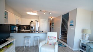 A modern living room with a TV, white armchair, and a kitchen area in the background. - Oceanfront 3BR townhome in Surf City with stunning views at Turtle Cove 908D. (Surf City)