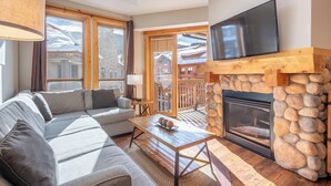 A cozy living room with a stone fireplace, a flat-screen TV mounted above it, and a large window offering a view of a snowy mountain.