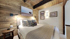A bedroom with a wooden ceiling, a bed with multiple pillows, a bedside table, and wall-mounted lights.
