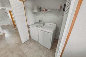 A laundry room with a washing machine and dryer, a shelf with cleaning supplies, and a door leading to another room.