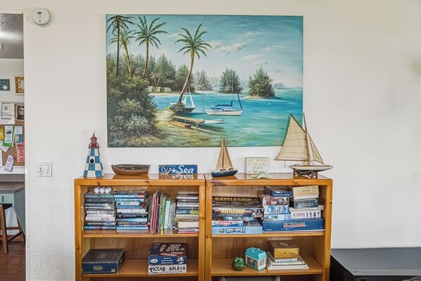 A bookshelf with various books and board games, a painting of a beach scene, and a small lighthouse model.