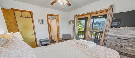 A bedroom with a bed, a desk, a chair, a ceiling fan, and a view of the outside.