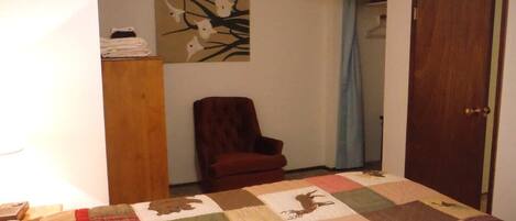 A bedroom with a quilted bedspread, a wooden chair, a door, and a wall with a painting.