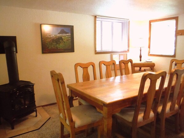 A dining area with a wooden table and chairs, a black stove, a framed picture on the wall, and a window with blinds.