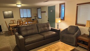Ein Wohnzimmer mit einer Couch, Sesseln, einem Couchtisch, einem Esstisch, Stühlen, Lampen und einer Tür.