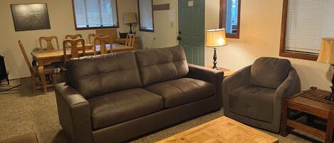 A living room with a sofa, armchair, coffee table, dining table, chairs, lamps, and a door.