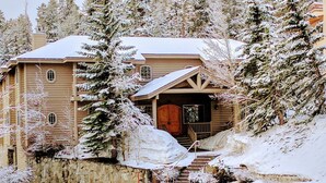 A two-story log cabin with a stone foundation, a wooden front porch, and a snow-covered landscape.