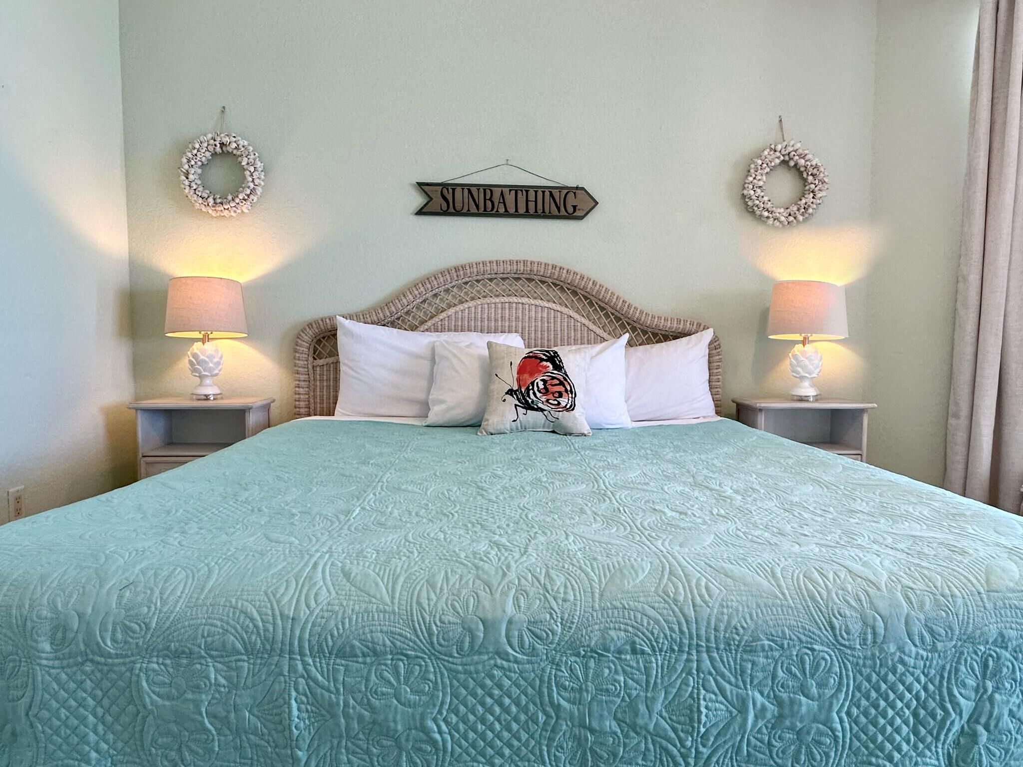 Un letto con una coperta azzurra, due cuscini bianchi e un cuscino decorativo con un motivo rosso e nero, con un cartello sopra il letto che dice "SUNBATHING".