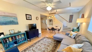 A living room with a sofa, a coffee table, a bookshelf, a TV, and a staircase leading to another room. - Off The Beaten Path:  Surrounded by trees and nicely secluded. (Ocracoke)