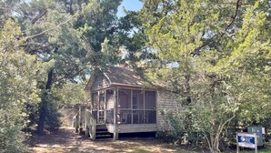 A small wooden cabin with a porch, surrounded by trees and a sign indicating it is for rental. - Off The Beaten Path:  Surrounded by trees and nicely secluded. (Ocracoke)