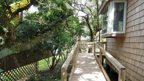 A wooden deck leading to a building with a window, surrounded by greenery and a fence. - Off The Beaten Path:  Surrounded by trees and nicely secluded. (Ocracoke)