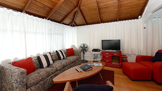 Uma sala de estar com teto de madeira, um sofá, uma poltrona vermelha, uma televisão e um ventilador.