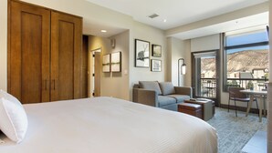 A hotel room with a large bed, a sofa, a balcony with a view, and a dining area.