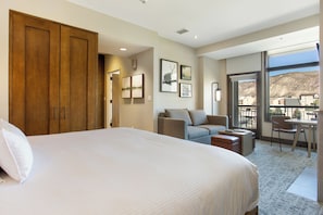 A hotel room with a large bed, a sofa, a balcony with a view, and a dining area.