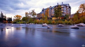 A modern hotel with a river in front, surrounded by autumn trees and a mountainous backdrop. - CRBC Remodeled 2024 Resort & Spa, Ski in out, Ski Valet, Health Club, Hot Tubs (Avon)