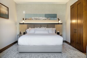 Ett modernt hotellrum med en stor säng, två sängbord, ett litet bord, en garderob i trä och en inramad landskapsmålning ovanför sängen.