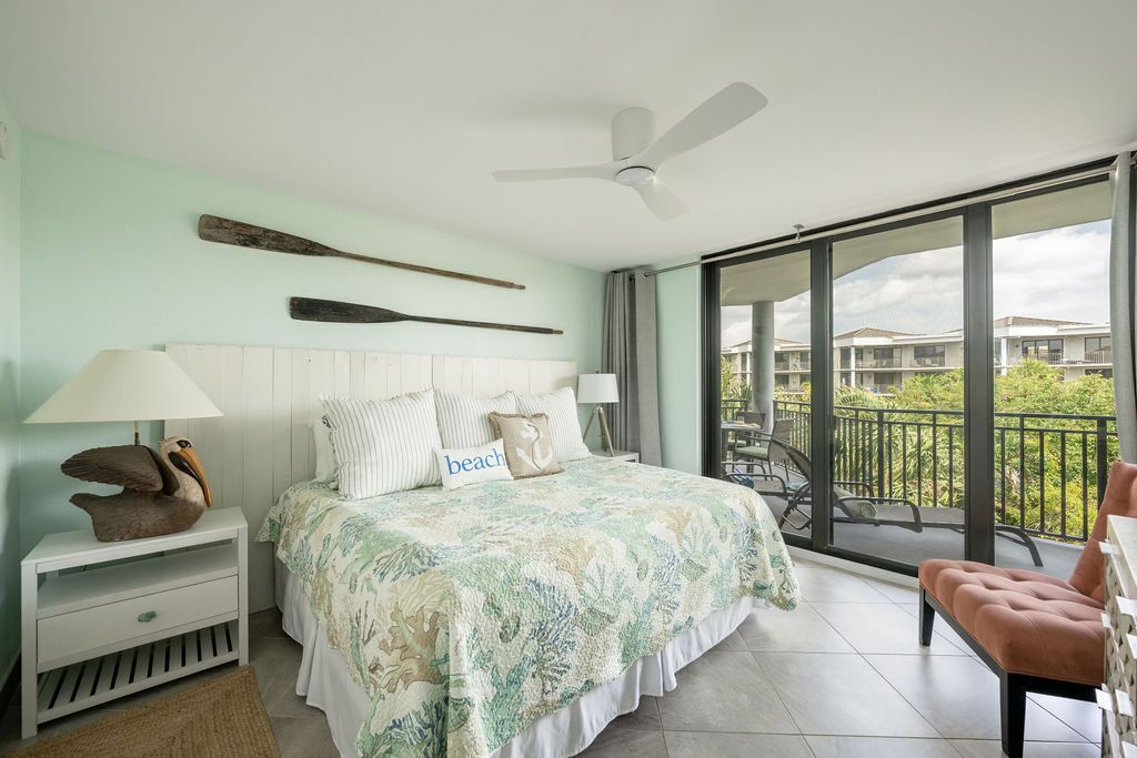 Een slaapkamer met een bed, nachtkastje, lamp, ventilator en een balkon met een stoel.