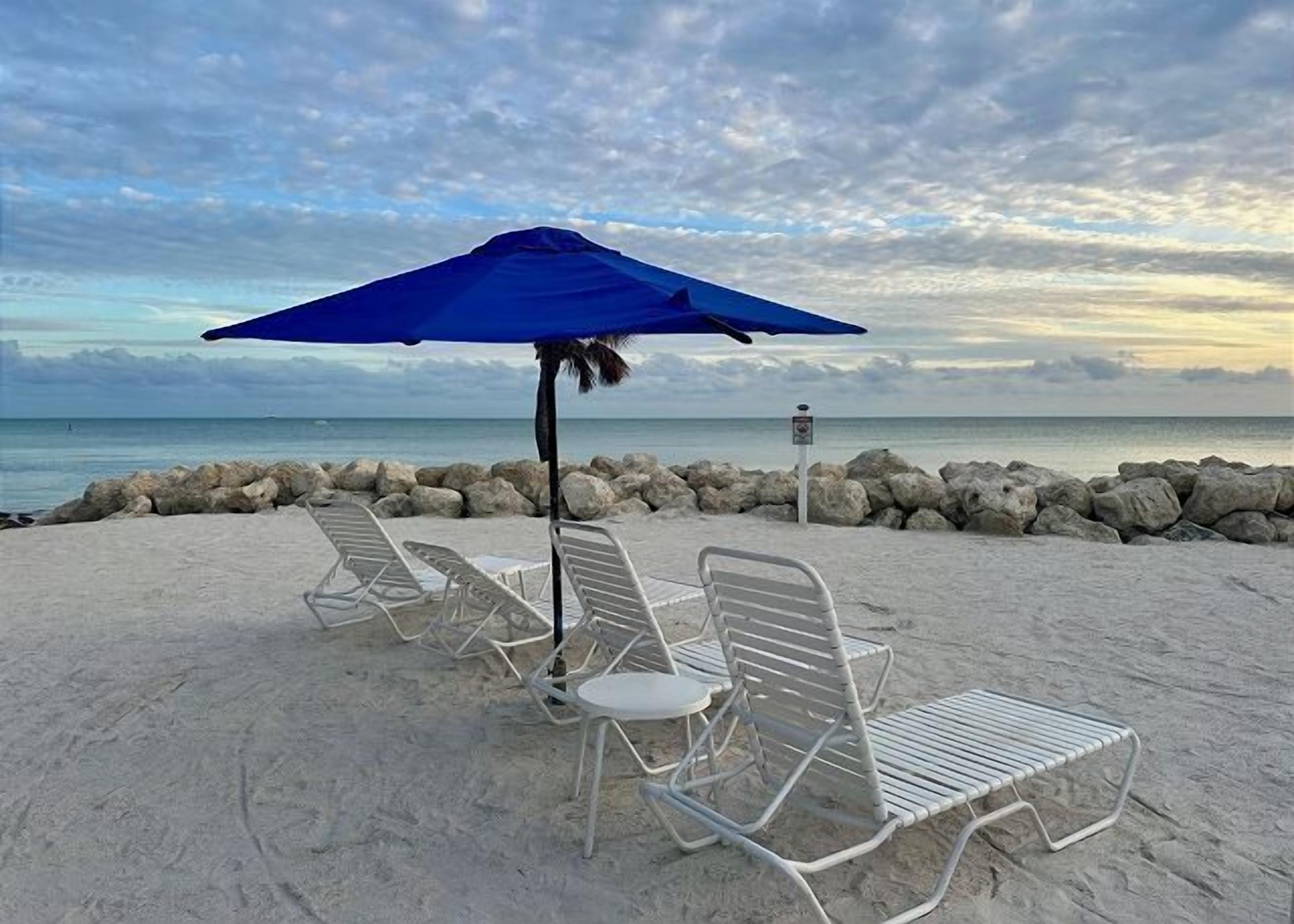 A beach with white metal lounge chairs and a blue umbrella.