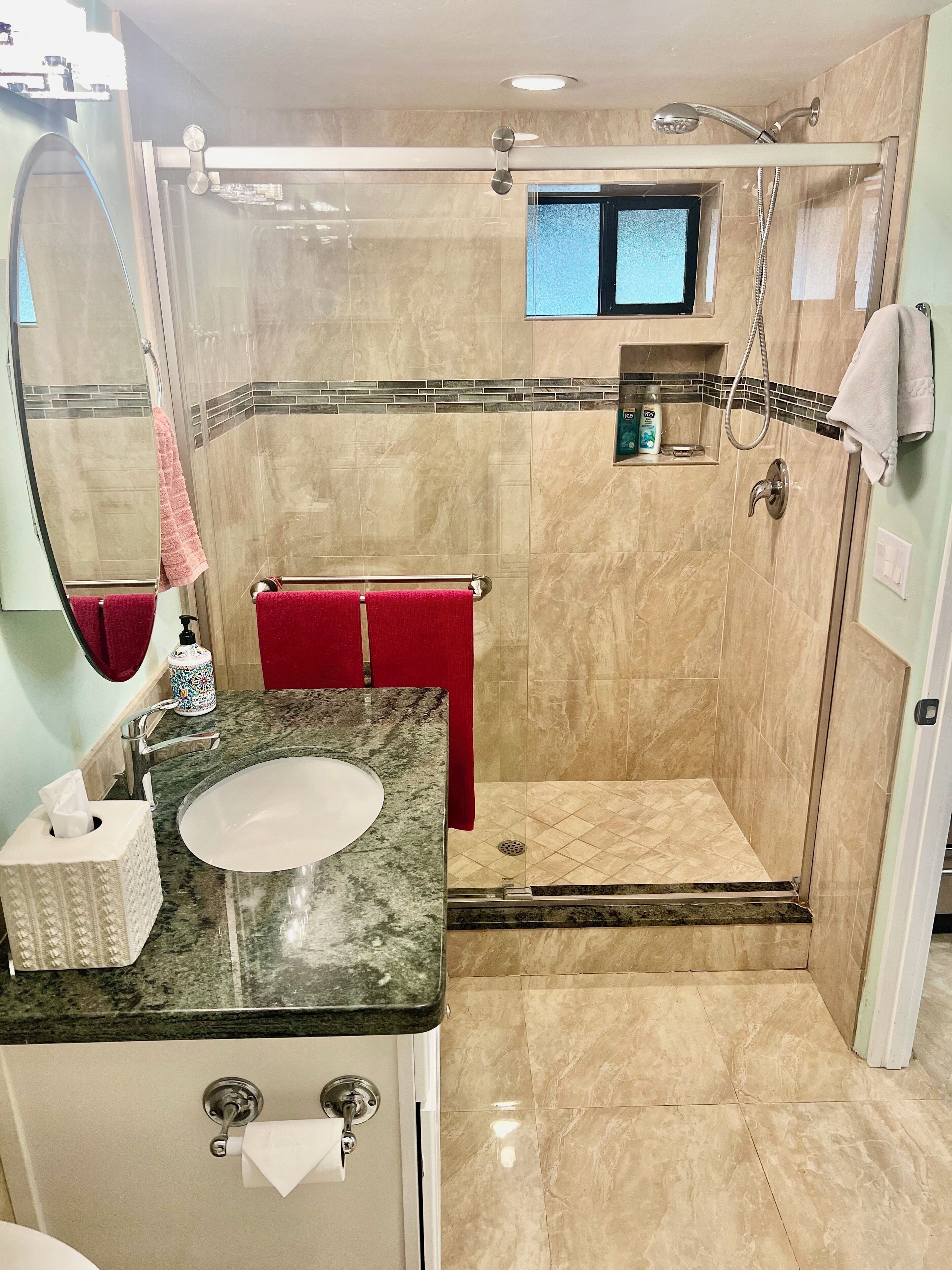 A bathroom with a sink, mirror, red towel, shower, and tiled walls.
