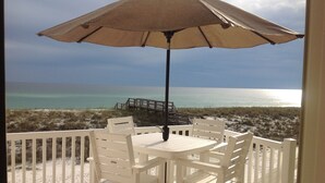 Un espace extérieur aménagé avec des meubles blancs, comprenant une table et des chaises, un parasol, et offrant une vue sur la plage et l’océan.