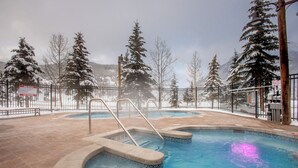 Uma área de piscina com hidromassagem, rodeada por árvores cobertas de neve e um cenário montanhoso.