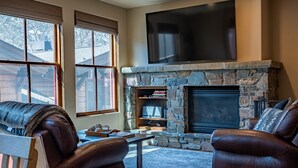 Una sala de estar acogedora con chimenea de piedra, sofás de cuero, un televisor de pantalla plana y ventanas grandes.