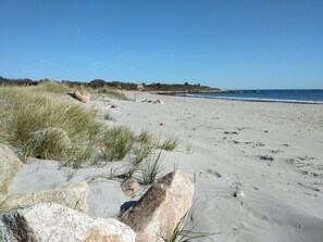 A sandy beach with rocks and grass in the foreground, a clear blue sky, and a calm sea in the background.