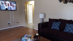A living room with a dark brown sofa, patterned cushions, a coffee table with magazines, a TV mounted on the wall, and a lamp on a side table. - A BEAUTIFUL 3 BEDROOM BUNGALOW WITH ALL MODERN AMENITIES  CLOSE TO A PARK!! (Dayton)