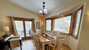 Un comedor con una mesa y sillas de madera, una araña y ventanas amplias con persianas.