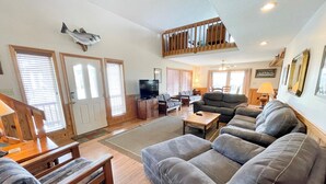 A spacious living room with a large grey sofa, a wooden bench, and a television on a stand. There is a staircase with a wooden railing leading to an upper level. - Blind Mullet:  Family friendly, den, decks, deep water private dock. (Ocracoke)